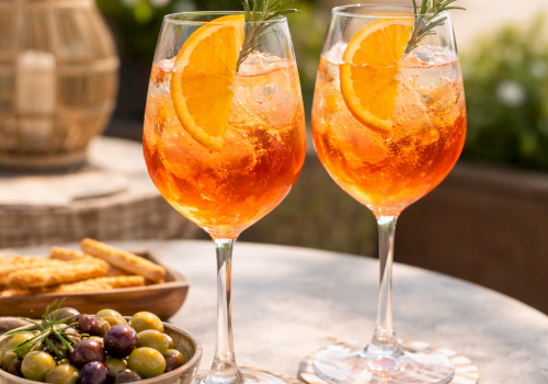 Two orange cocktails with orange slices and rosemary on a sunny outdoor table, plus a bowl of olives and breadsticks in the scene.