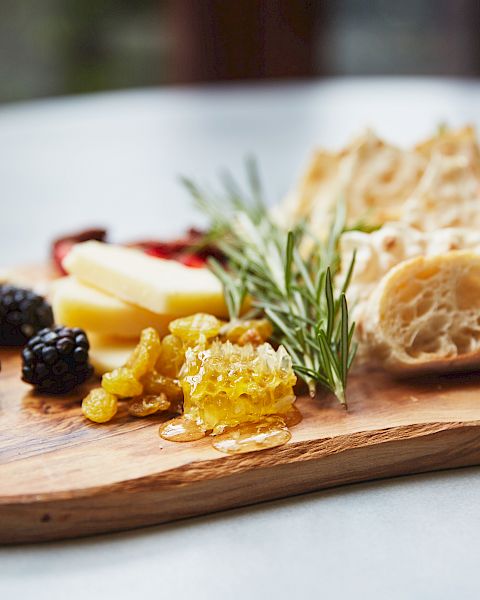 A cheese board with assorted items: cheeses, bread, olives, jam/honey, herbs, and cured meats on a wooden serving board.