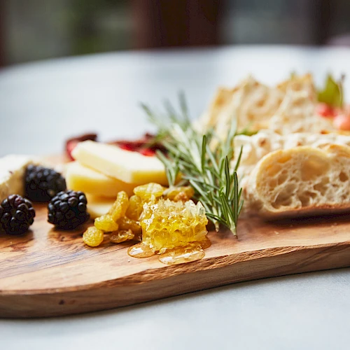 A cheese board with assorted items: cheeses, bread, olives, jam/honey, herbs, and cured meats on a wooden serving board.
