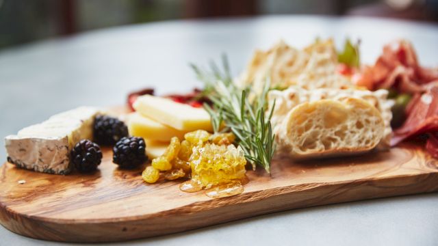 A cheese board with assorted items: cheeses, bread, olives, jam/honey, herbs, and cured meats on a wooden serving board.