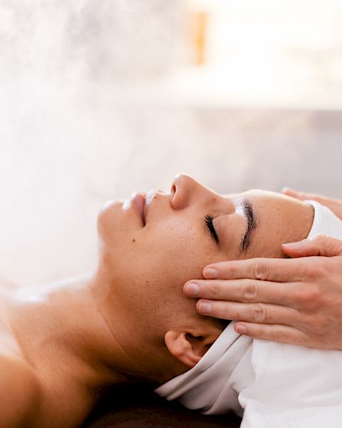 A woman receiving a steam facial or spa treatment with a therapist massaging temples, creating a relaxing wellness scene.
