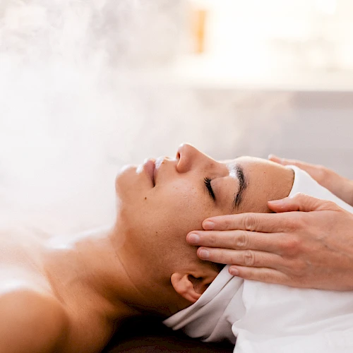 A woman receiving a steam facial or spa treatment with a therapist massaging temples, creating a relaxing wellness scene.