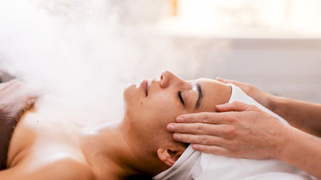 A woman receiving a steam facial or spa treatment with a therapist massaging temples, creating a relaxing wellness scene.