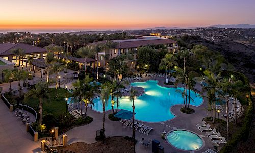 A resort pool complex at dusk with multiple pools, lounge chairs, palm trees, and surrounding buildings, city lights in the distance, sunset glow.