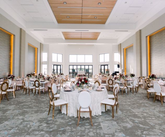 A spacious banquet hall with round tables, white tablecloths, pink floral centerpieces, and cushioned chairs set for a formal event, bright natural light.