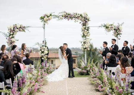 A couple stands at the end of an aisle under a floral arch, surrounded by guests in a garden setting, ready to wed.