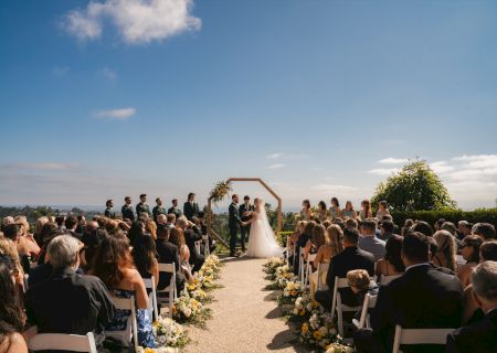 A wedding ceremony outdoors with a bride and groom at the altar, surrounded by guests seated on both sides along an aisle, under a bright blue sky.
