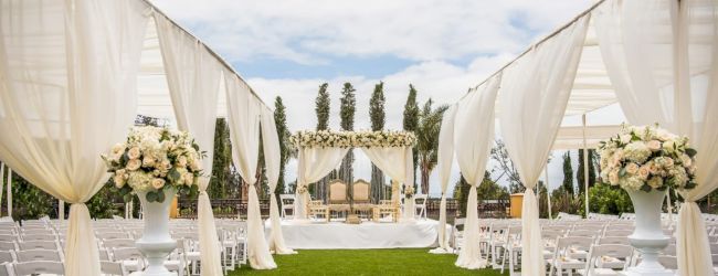 An elegant outdoor wedding setup with white draped arches, floral arrangements, and rows of chairs on a green lawn under a bright sky.