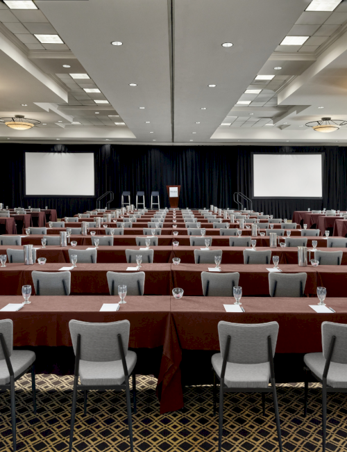 A large conference room set for a formal event with rows of tables and chairs facing a stage, two projectors, and a lectern.