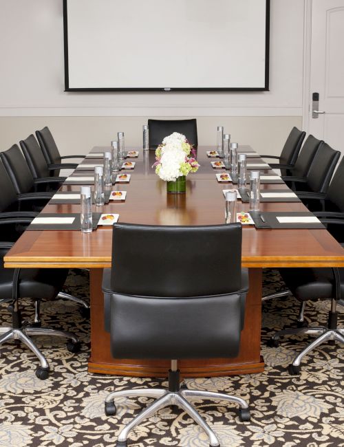A long conference table with black rolling chairs, place settings, and a floral centerpiece in a formal boardroom.