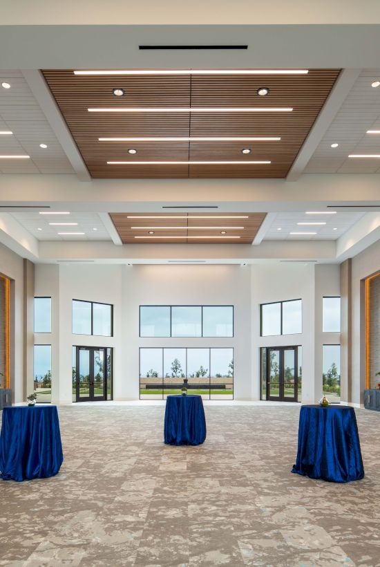 A spacious event hall with high ceilings, recessed lighting, and three round tables draped in blue cloths; windows reveal a bright, airy view.