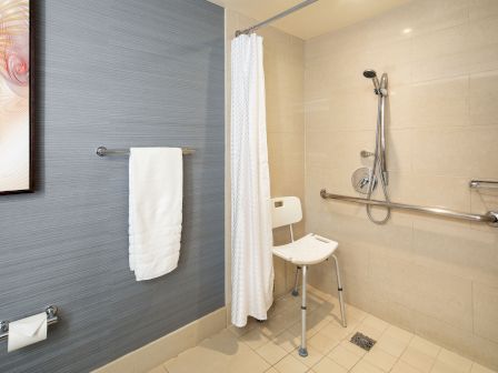 A modern accessible hotel bathroom with a shower chair, handheld shower, grab bars, a white curtain, towel rack, and beige tiled walls and floor.