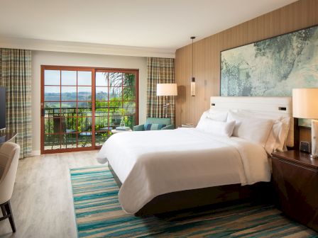 A modern hotel room with a large bed, nightstands, a flat screen tv, a balcony view, a rug, and soothing wall art by the headboard, ending the sentence.