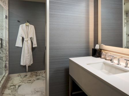 A modern hotel bathroom with a white bathrobe hung on the door, a glass shower, marble floor, and a wide sink counter with a mirror and lighting, end.