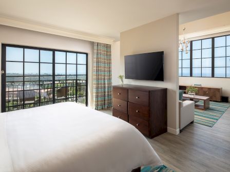 Modern hotel room with a large bed, dresser, mounted TV, and a bright living area; big windows offer city views and airy decor.