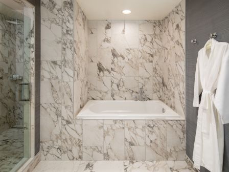 A marble-tiled bathroom with a built-in bathtub, glass shower to the left, and a white robe hanging on a hook to the right.