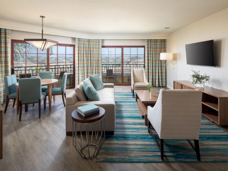 A bright hotel suite with a living room and dining area: two sofas, armchairs, coffee table, turquoise rug, wooden cabinets, a wall-mounted TV, and large windows with plaid curtains.