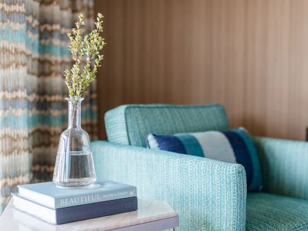A glass vase with yellow-green flowers sits on a stack of books beside a teal sofa, creating a bright, cozy living room corner.