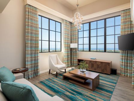 A modern living room with large grid windows, colorful plaid curtains, a teal rug, white sofa and chair, wooden coffee table, and a wall-mounted TV.