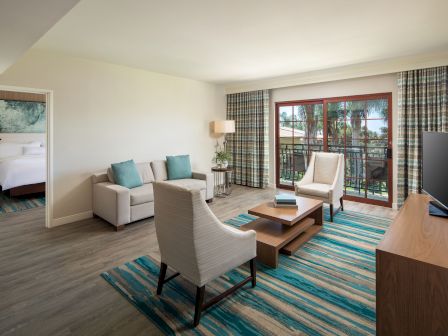 A modern hotel suite living area with a sofa, two armchairs, a coffee table, a rug, TV on a console, and sliding glass doors opening to a balcony, with a doorway to a bedroom.