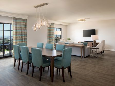 A bright, open living-dining area with a long table and teal chairs, a kitchen island, a sofa, wall TV, and large windows with curtains. End.