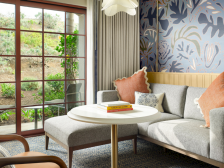 A bright, cozy living room with a gray sectional, peach cushions, and a round coffee table; sliding glass doors open to a green garden.