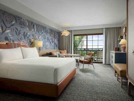 A stylish hotel room with a large bed, floral wallpaper, seating area, desk, and balcony view.