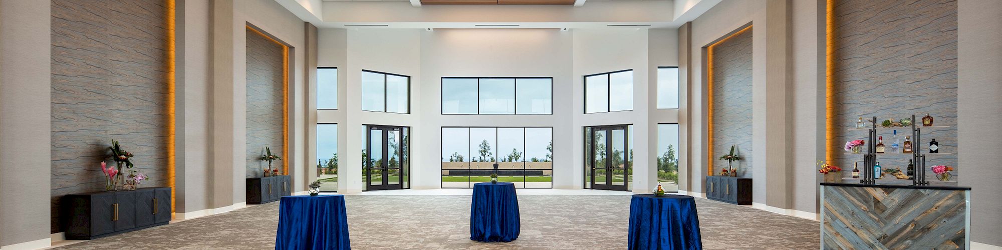 A spacious, well-lit conference hall with high ceilings, modern recessed lighting, three round cocktail tables draped in blue cloth, and large windows at the far end.