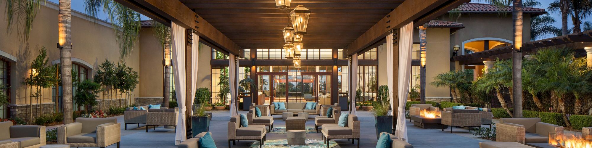 A stylish outdoor lounge area with cushioned seating, low tables, and hanging lanterns under a wooden pergola, arranged for communal relaxation.