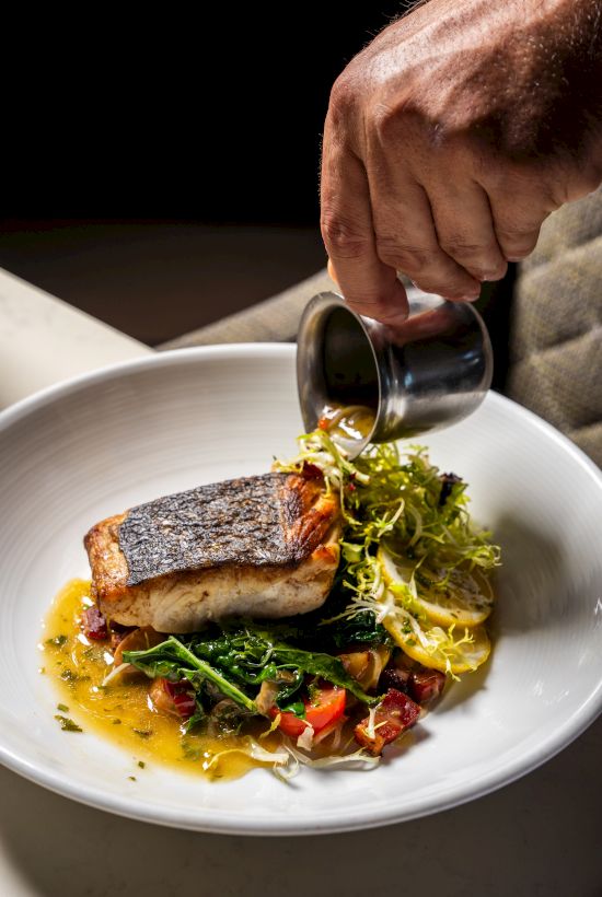 Chef pours sauce over a plated fish dish with greens, vegetables, and a crisp crust, on a white plate.