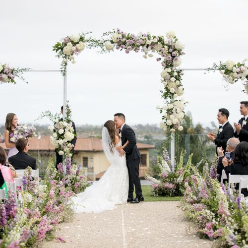 A wedding ceremony outdoors: couple kisses under a floral arch with guests seated along a flower-lined aisle. All on a sunny day.