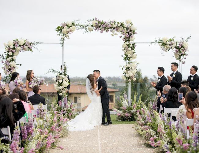 A wedding ceremony outdoors: couple kisses under a floral arch with guests seated along a flower-lined aisle. All on a sunny day.