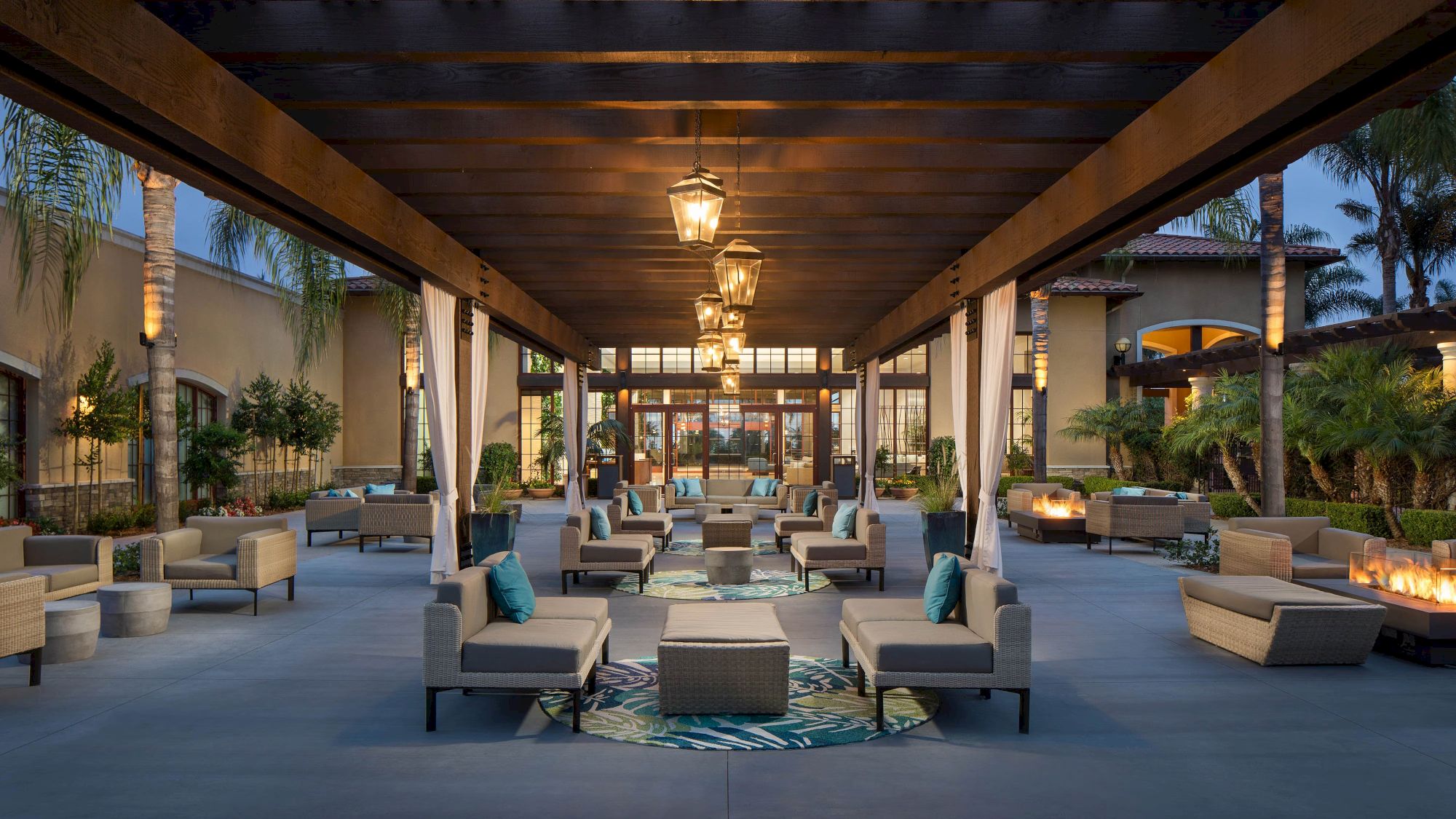 A stylish outdoor lounge area with couches, coffee tables, and warm lighting under a wooden pergola, surrounded by greenery and fire features.