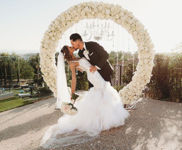 A bride and groom kiss in front of a large flower heart arch at an outdoor wedding, with a scenic backdrop and soft sunlight.