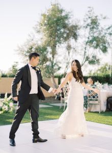 A bride in a white dress and a groom in a black suit hold hands and dance outdoors at a wedding reception.