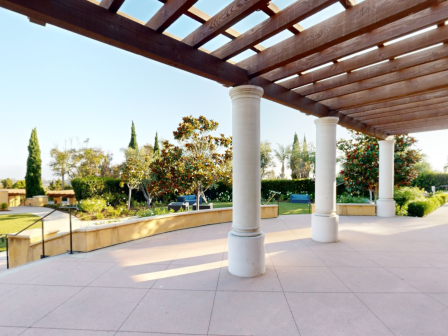A sunny outdoor scene under a wooden pergola with white columns, a paved patio, manicured greenery, and tidy park-like landscaping.