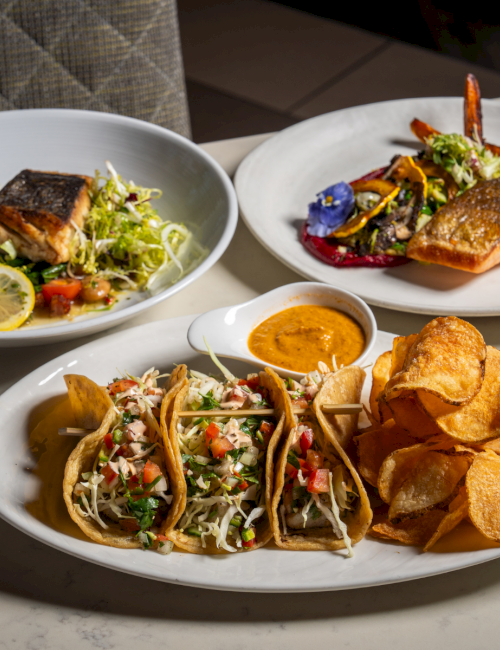 Three plated dishes: salmon with greens, another fish with veggies, and tacos with salsa and chips, all served on white plates.