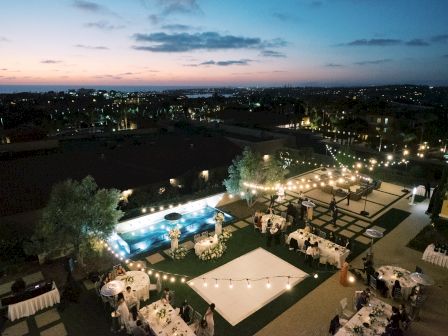 Nighttime outdoor wedding or event setup with string lights, round tables, tents, and a pool area overlooking a city skyline at dusk.