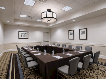 A conference room with a U-shaped table setup, grey chairs, water bottles, notepads, framed photos on the walls, and ceiling lights.