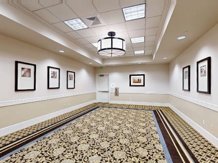 A carpeted hallway with framed pictures on cream walls, ceiling lights, and a decorative central chandelier, empty and well-lit.