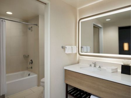 A modern hotel bathroom: vanity with double basin and large backlit mirror, towels on rack, and a separate shower-tub combo.