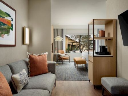 A cozy hotel suite with a gray sofa and orange pillows, a wooden cabinet, and a view into a bright living area with a rug, chairs, and a large window.
