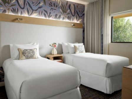 Two neatly made hotel beds with white linens, a shared nightstand, flower vase, patterned headboard, and a window with curtains.
