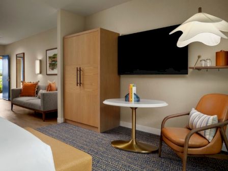A modern hotel suite with a round white table, orange accent chair, wall-mounted TV, wooden cabinet, and a gray-blue patterned carpet, cozy seating area.