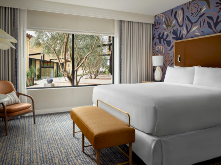 A stylish hotel room with a large window view, king bed, mustard bench, blue patterned wall, chairs, and bright natural light.