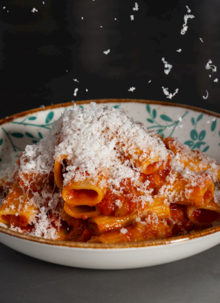 A bowl of pasta tossed in tomato sauce, topped with shredded cheese, on a decorative plate with a dark background.
