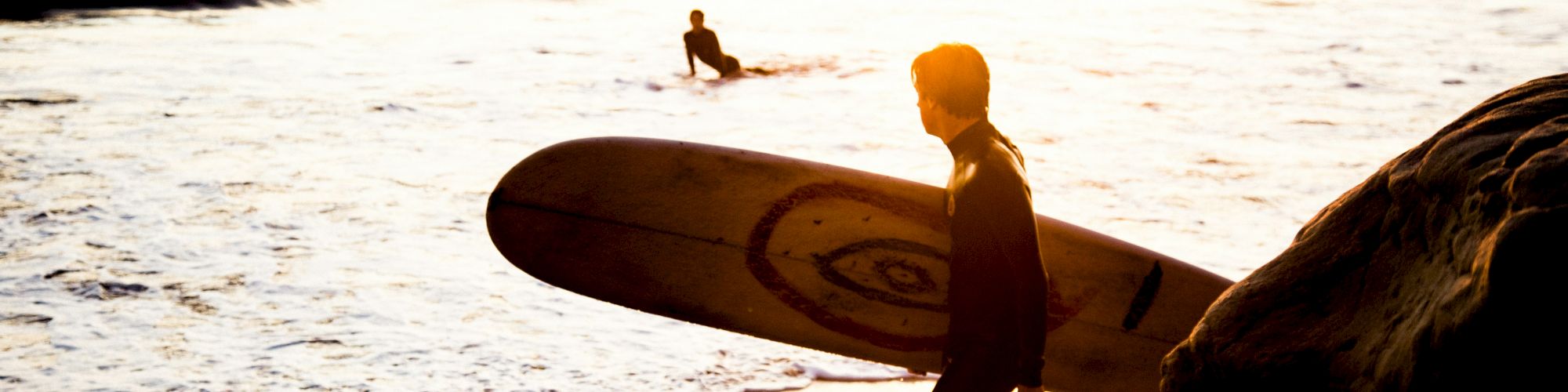 A person carrying a surfboard on the beach at sunset, about to enter the waves, with another surfer in the water nearby.