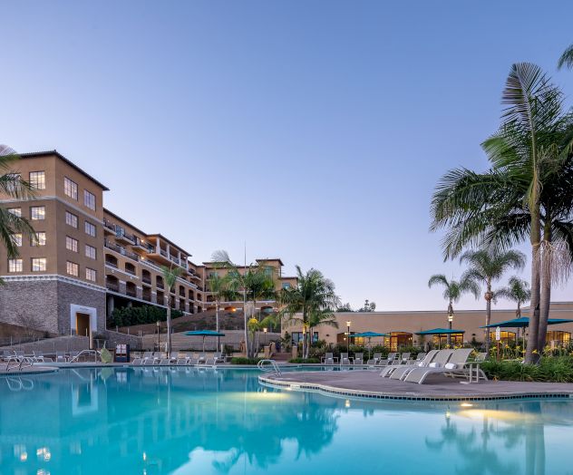 A resort-style hotel with a large outdoor swimming pool, palm trees, and multiple buildings in the background, under a clear sky.