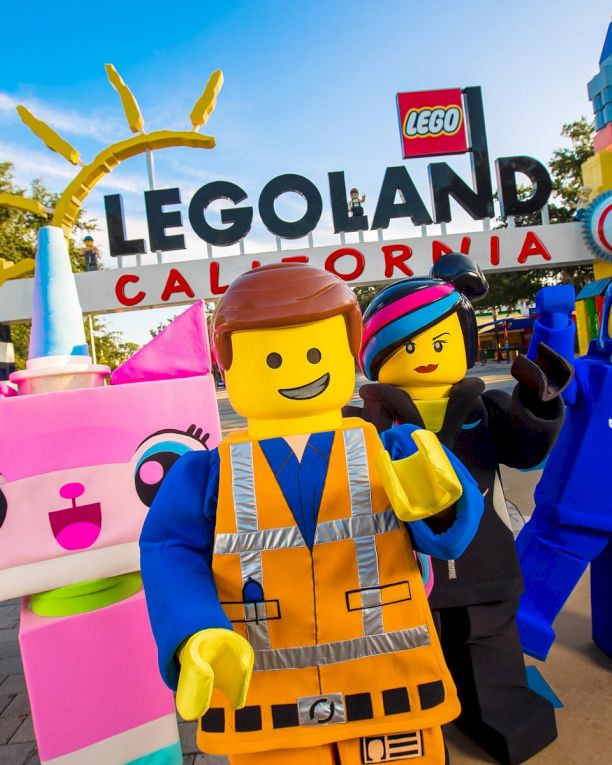 A group of LEGO characters posing in front of Legoland California, with a bright blue sky and colorful structures in the background. (140 chars max, ending with a period)