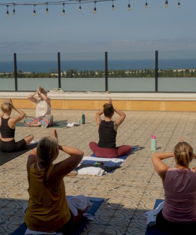 Outdoor yoga session on a rooftop terrace by the sea; people sit on mats, practicing poses as a guided instructor leads.